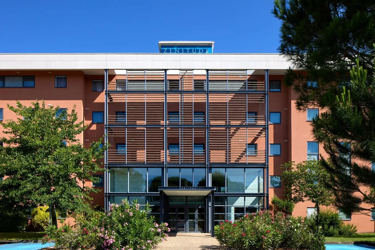 Façade moderne de l'hôtel Zenitude Bordeaux Aéroport avec murs terracotta, panneaux de brise-soleil horizontaux, entrée vitrée et aménagement paysager