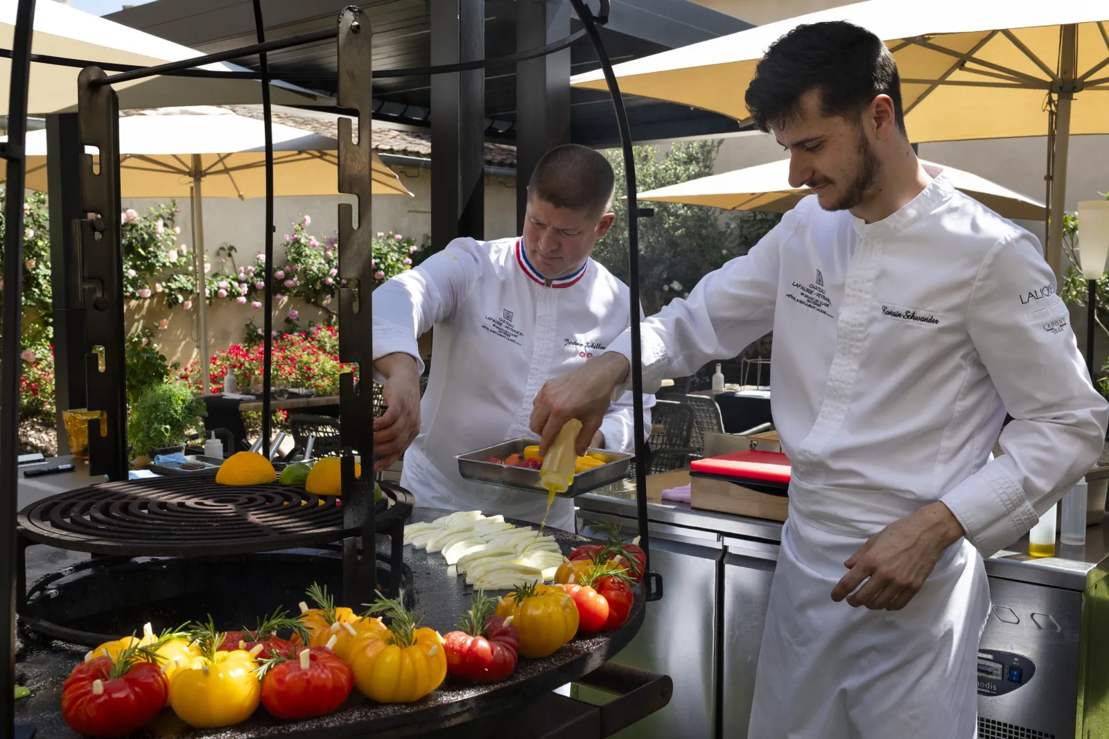 Deux chefs cuisiniers en tenue blanche professionnelle préparent des légumes frais (tomates et courgettes) sur une terrasse ombragée avec parasols jaunes à Bordeaux, La Terrasse de Lafaurie.