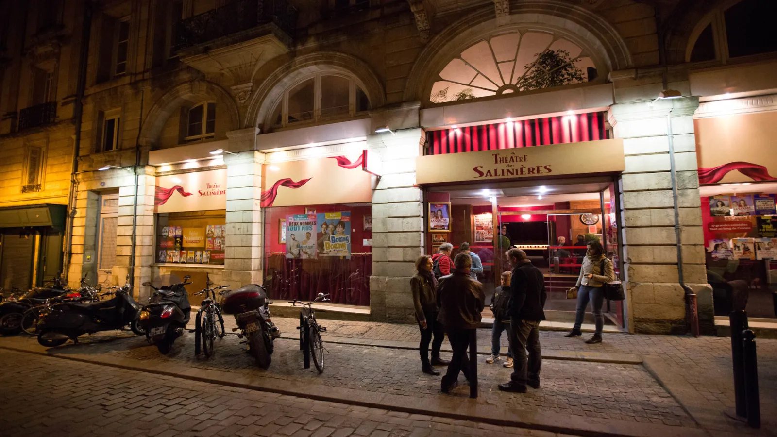 Façade du Théâtre des Salinières à Bordeaux illuminée la nuit, avec son enseigne rouge caractéristique et des spectateurs devant l'entrée