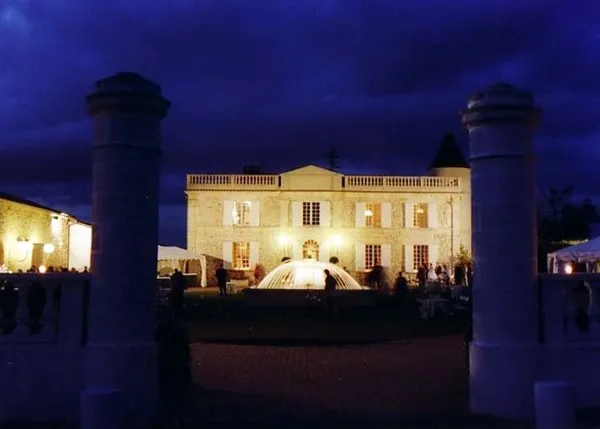 Vue nocturne du Château Lafitte à Bordeaux, illuminé en jaune, avec ses façades classiques encadrées par deux colonnes cylindriques et une fontaine en avant-plan, sous un ciel crépusculaire bleu profond.