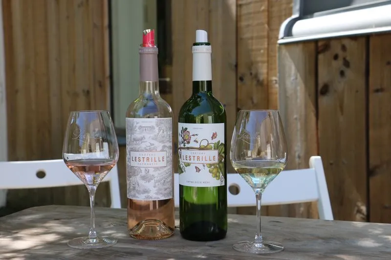 Deux verres de vin du Château Lestrille, l'un contenant un rosé et l'autre un blanc sec, accompagnés de deux bouteilles de vin sur une table en bois avec chaises blanches en arrière-plan.
