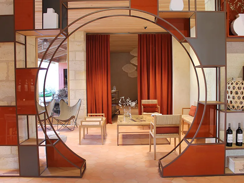 Exposition intérieure du Château Marquis d'Alesme Becker à Bordeaux montrant un salon moderne avec mobilier design, rideaux rouges, arche métallique arquée, et étagères géométriques en bois et rouge.