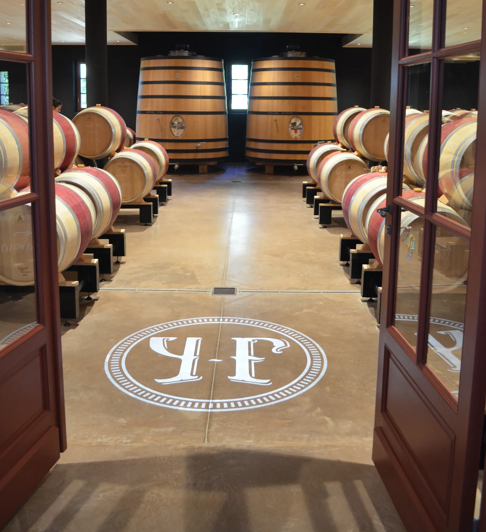 Cave de vinification du Château Yon-Figeac à Bordeaux avec des rangées de fûts de chêne alignés de chaque côté et deux grands foudres au fond, sol en béton poli avec le logo YF au centre