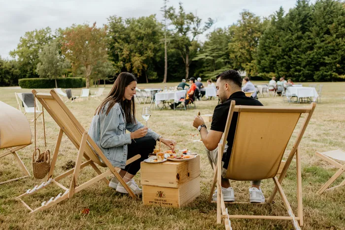 Deux personnes assises dans des transats en bois partagent un repas sur une plage aménagée à Bordeaux Afterbeach en Médoc, avec d'autres visiteurs et parasols en arrière-plan dans un cadre de détente estival.