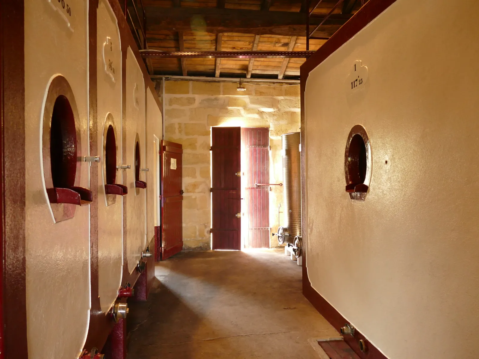 Couloir de cave viticole avec portes de tonneaux en bois rouge et blanc, lumière naturelle entrant par l'entrée au fond, Château Laniote à Bordeaux