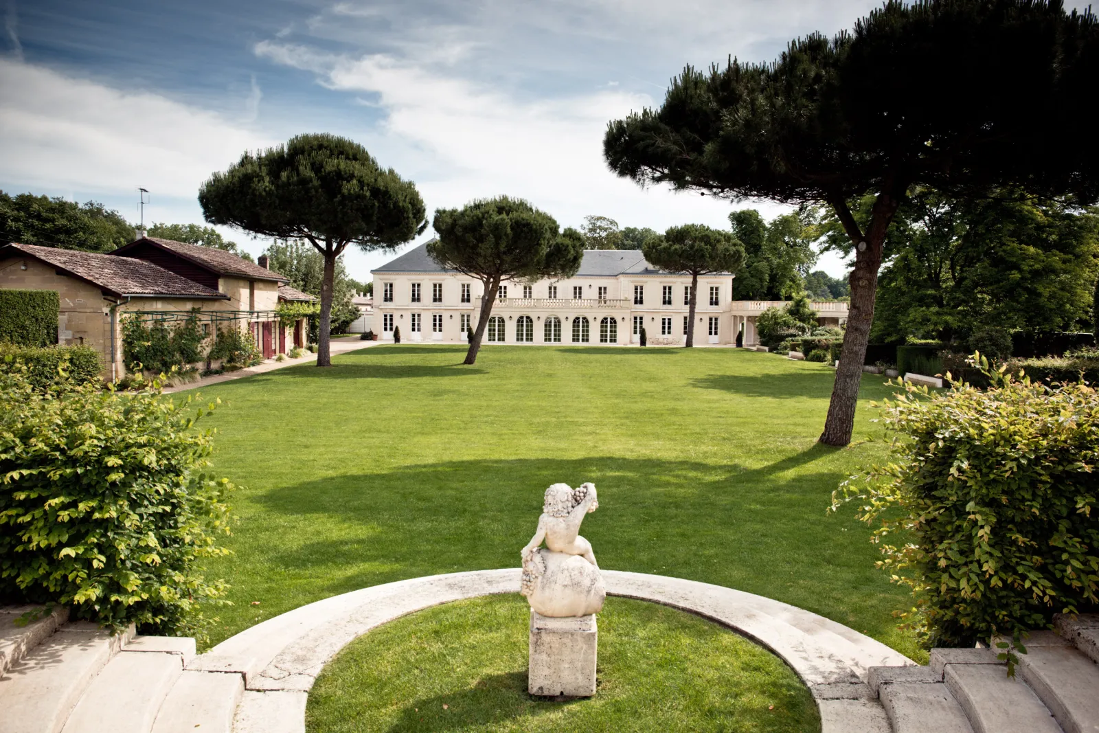 Vue d'ensemble du Château Malartic-Lagravière à Bordeaux, avec sa façade classique en pierre blanche, entouré d'une vaste pelouse verte, de pins parasols et de jardins paysagés, avec une sculpture en pierre au premier plan.