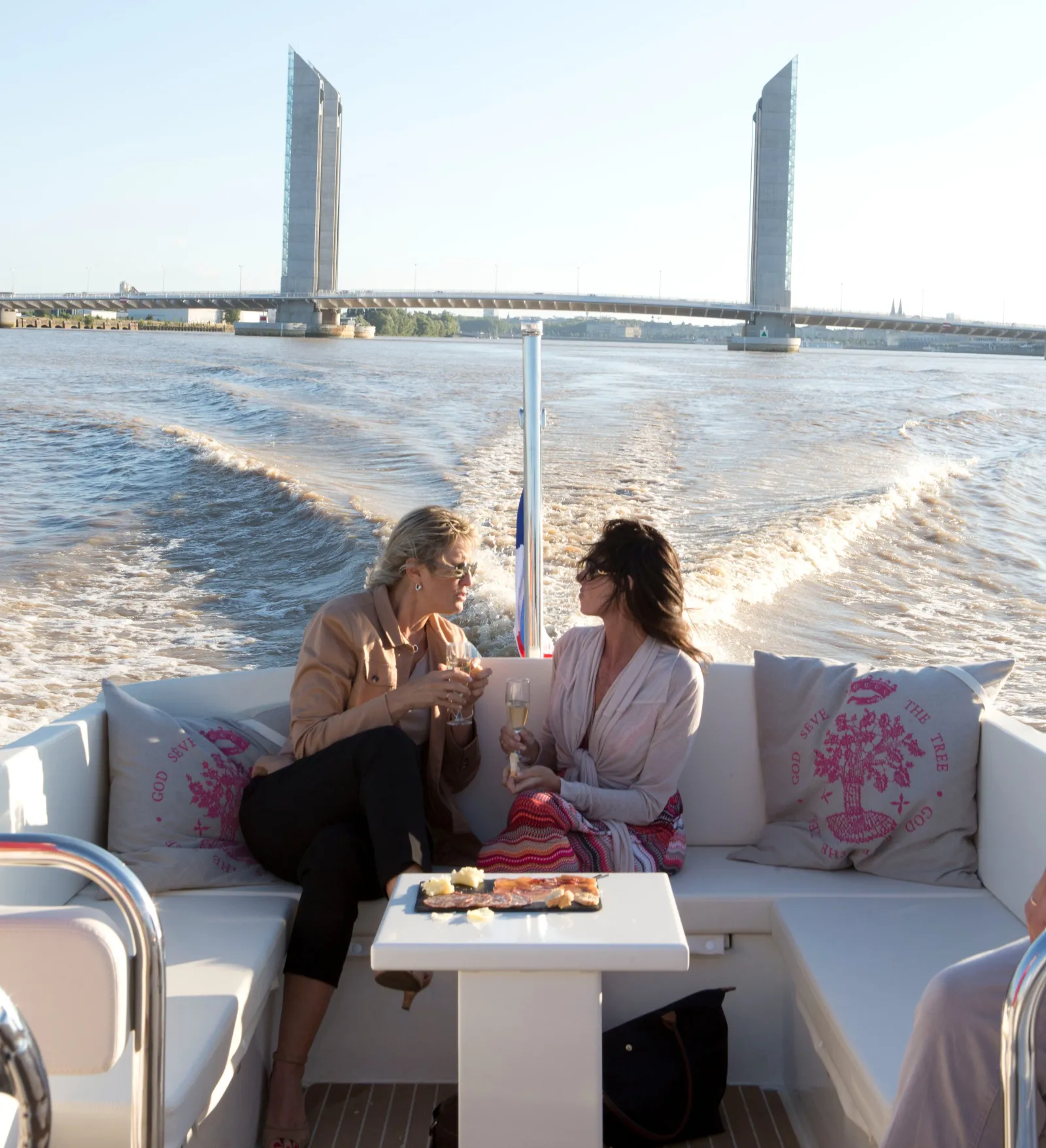 Deux femmes assises sur un canapé blanc dans un bateau-taxi Grattequina sur la Garonne à Bordeaux, avec le pont de Pierre et ses deux tours caractéristiques en arrière-plan
