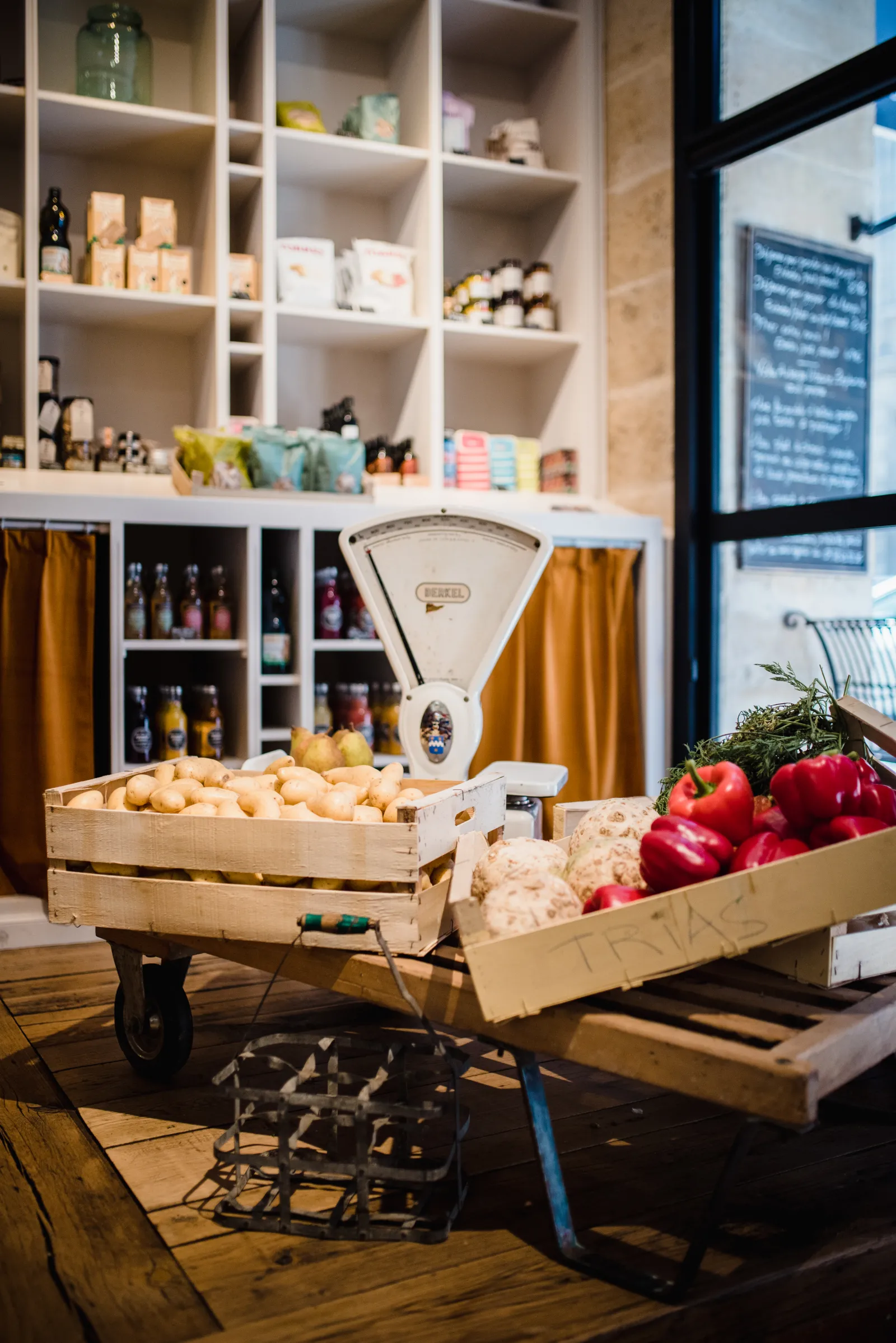 Intérieur d'une épicerie fine à Bordeaux avec deux caisses en bois contenant des légumes frais (oignons, poivrons rouges, chou-fleur), une balance vintage blanche au fond, et des étagères murales remplies de produits alimentaires et de bouteilles.