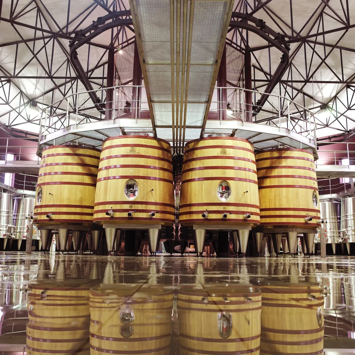 Rangée de grands fûts de chêne jaune et rouge alignés dans une cave de vinification moderne du Château Malartic-Lagravière à Bordeaux, avec reflet sur le sol brillant et structure métallique apparente au plafond.