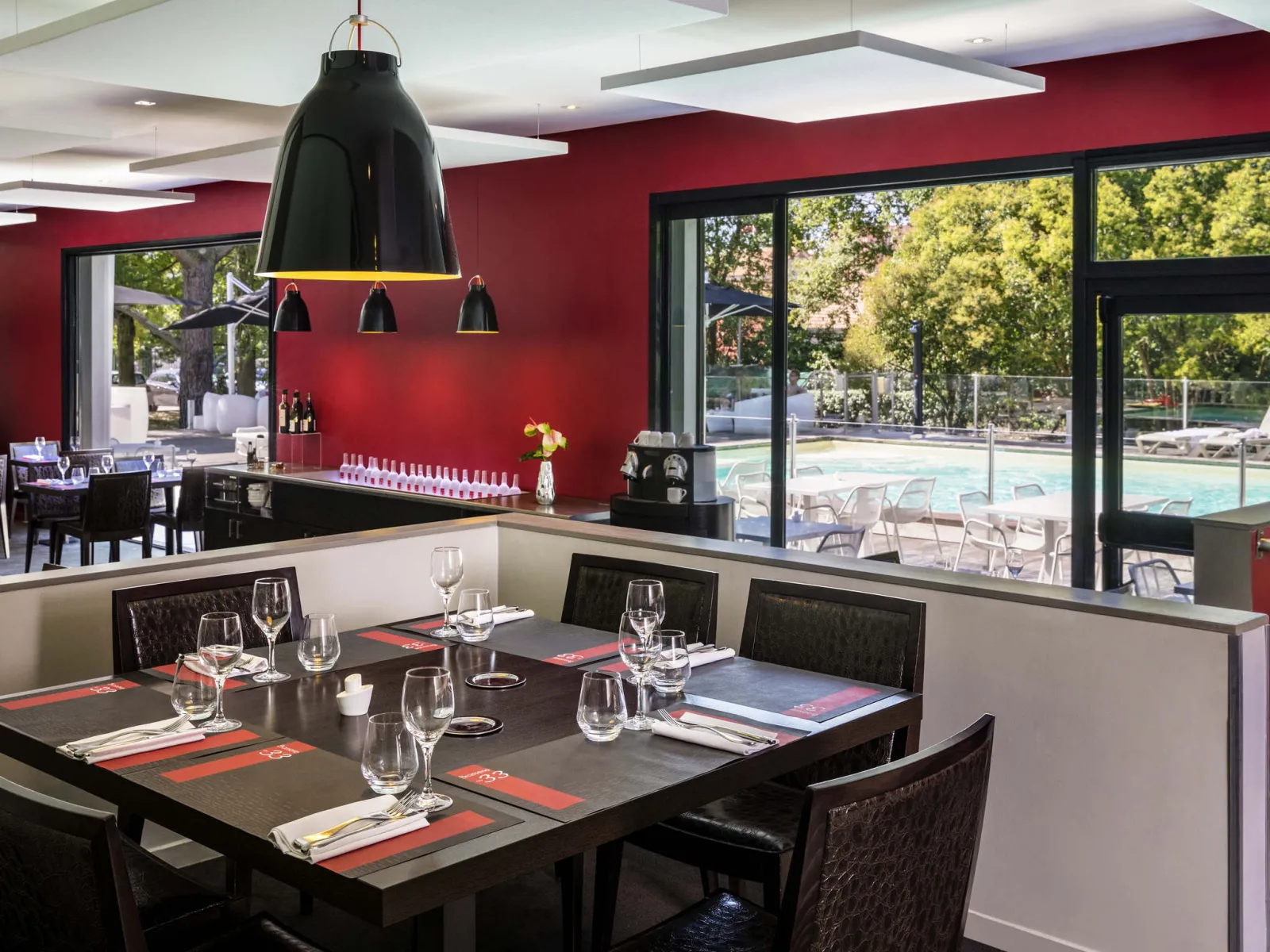 Salle de restaurant moderne avec murs rouges, tables noires dressées avec verres et couverts, bar noir avec suspension, grandes baies vitrées donnant sur une piscine et des transats en extérieur.