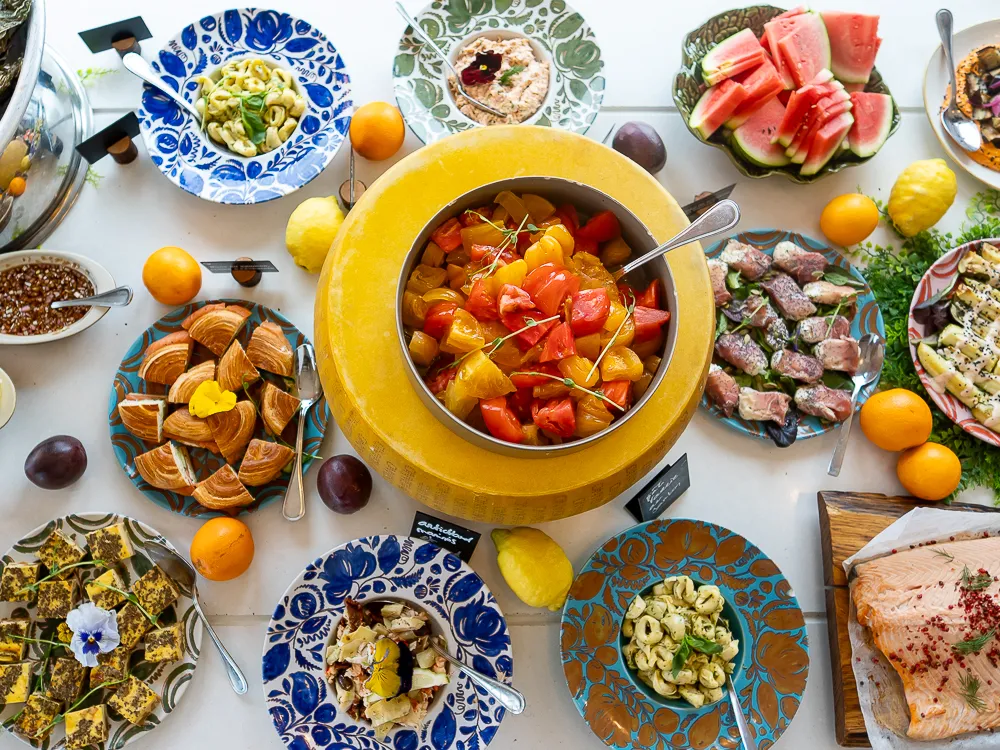 Table de buffet colorée avec un plat central de fruits rouges et jaunes, entouré de charcuterie, pastèque, croissants, fruits secs et diverses préparations culinaires sur vaisselle décorée.