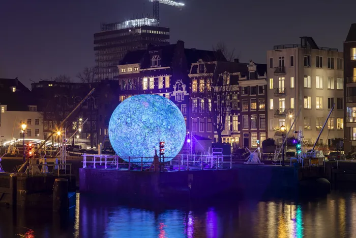 Installation artistique lumineuse représentant une sphère bleue géante illuminée sur les quais de Bordeaux la nuit, entourée de bâtiments historiques éclairés en violet, reflet dans l'eau du fleuve.