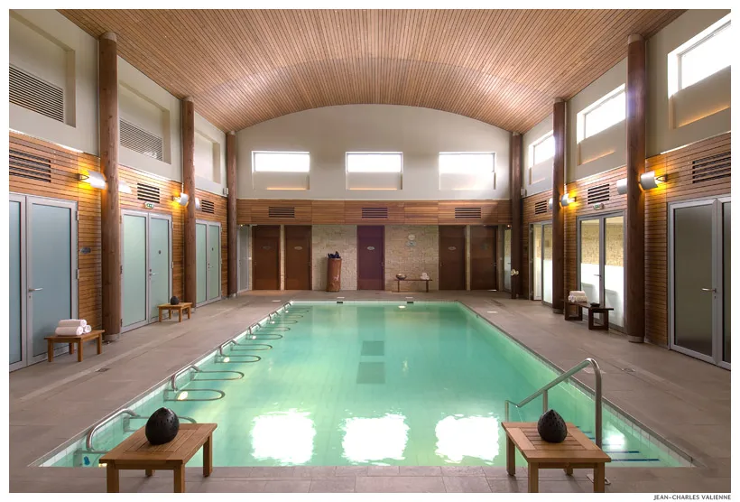 Piscine intérieure moderne du spa Les Bains de Margaux à Bordeaux, avec bassin rectangulaire aux eaux turquoise, entouré de cabines de change en bois, d'éclairages et d'une architecture contemporaine aux plafonds voûtés.