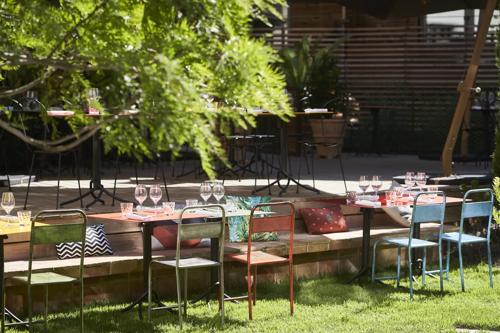 Terrasse extérieure de l'Hôtel La Zoologie à Bordeaux avec tables en bois, chaises colorées, verres à vin, végétation luxuriante et structures en bois ajouré