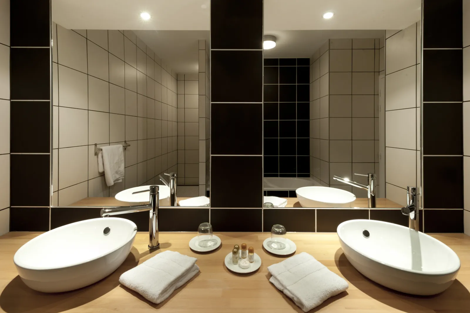 Salle de bain luxueuse du Château La Chèze à Bordeaux avec deux vasques blanches ovales sur plan de travail en bois, miroir panoramique, carrelage noir et beige, et accessoires de toilette haut de gamme