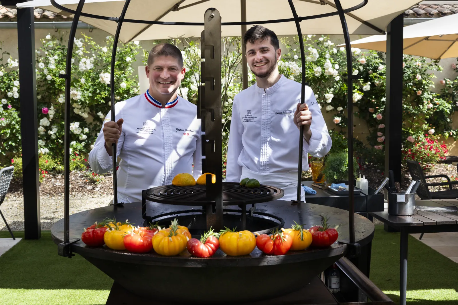Deux chefs cuisiniers en tenue blanche posent devant un brasero circulaire garni de tomates colorées, dans un jardin fleuri avec pergola à Bordeaux.