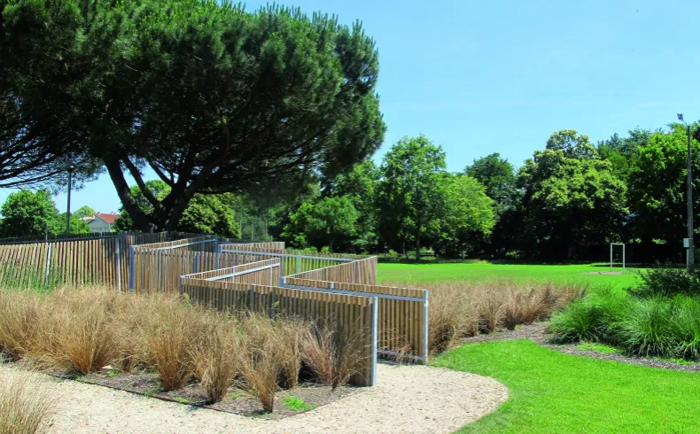 Parc aménagé aux bords de la Jalle de Gajac à Bordeaux avec pelouse verte, plantes ornementales, clôtures en bois et grands arbres sous un ciel bleu
