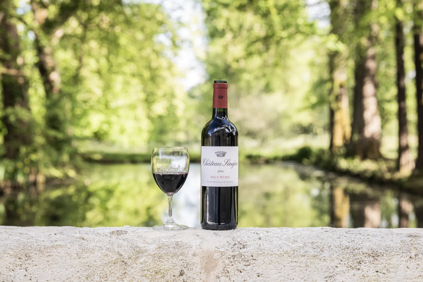 Bouteille de Château Sénéjac 2016 Haut-Médoc avec un verre de vin rouge rempli, posés sur une surface en pierre avec une allée d'arbres en arrière-plan.