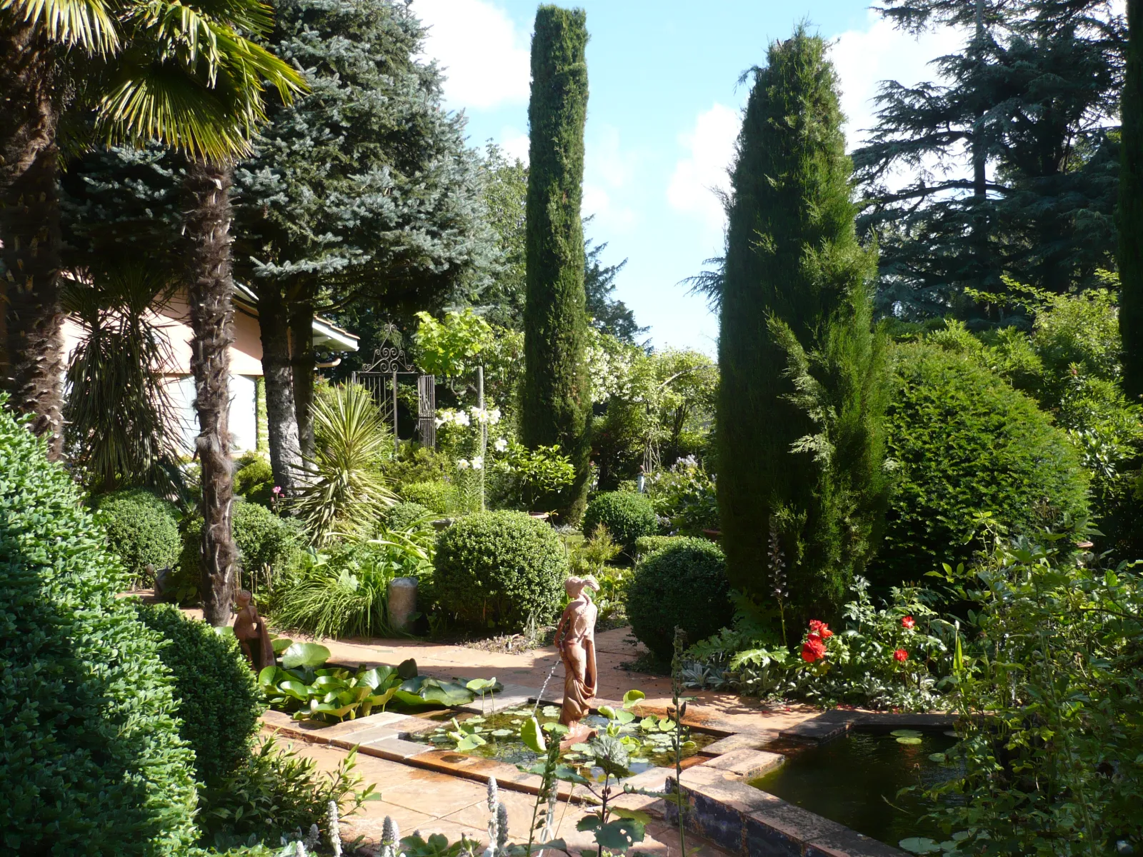 Vue du Jardin du Clozet à Bordeaux avec allée pavée, bassins d'eau, cyprès élancés, palmiers, sculptures et végétation luxuriante