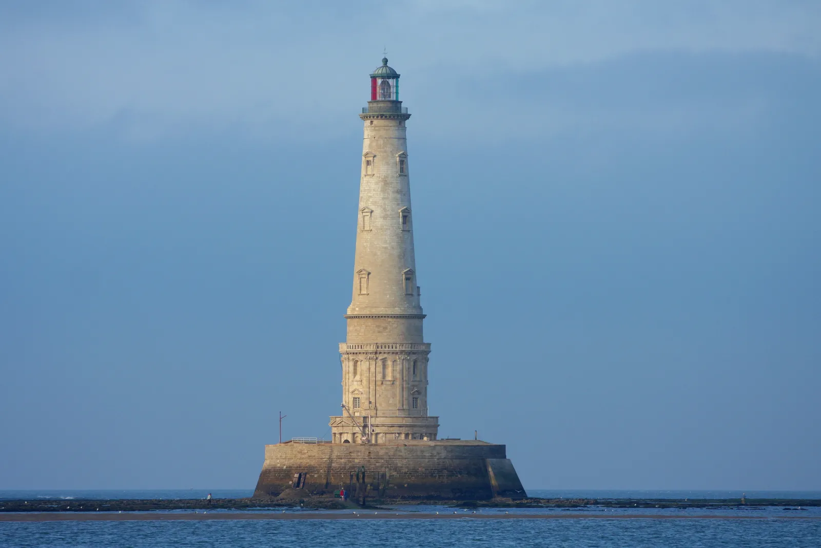 Le Phare de Cordouan, un phare historique situé sur un îlot rocheux en mer, au large de Bordeaux, avec sa tour blanche élancée et sa lanterne rouge au sommet.