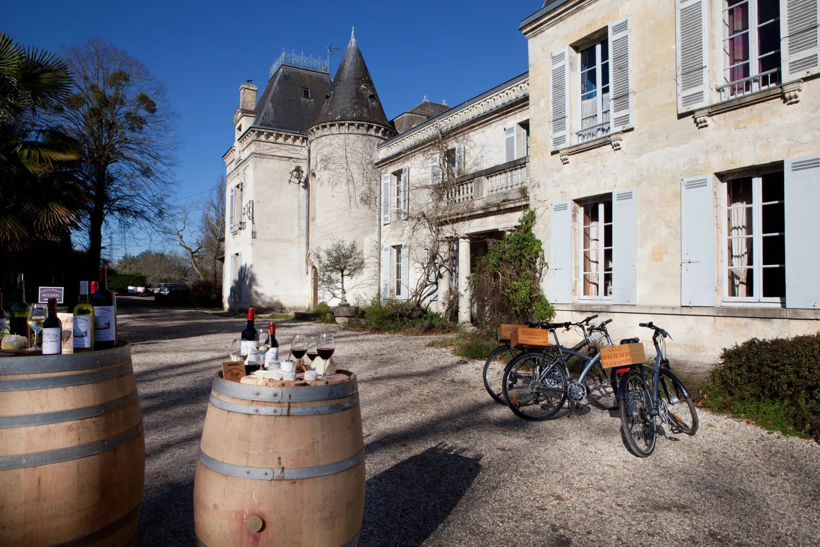 Cour d'un château avec tonneau de vin, bouteilles, verres, vélos de location et bâtiment historique aux tours pointues sous un ciel bleu