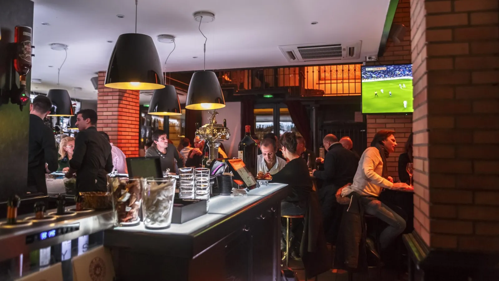 Intérieur animé du bar Bodega Bodega à Bordeaux avec clients au comptoir, luminaires noirs, briques rouges, match de football sur écran mural et verres remplis au premier plan.