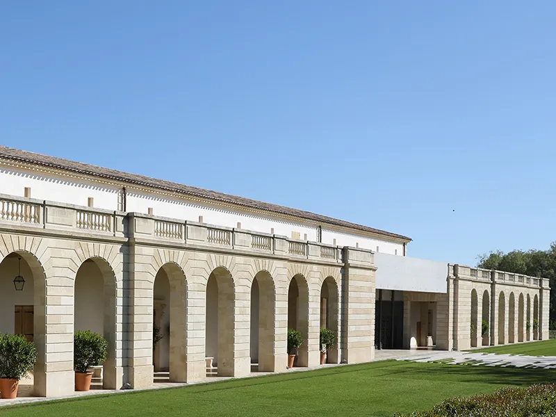 Façade du Château Marquis d'Alesme Becker à Bordeaux, architecture néoclassique avec colonnade de pierre, terrasse en herbe et ciel bleu.