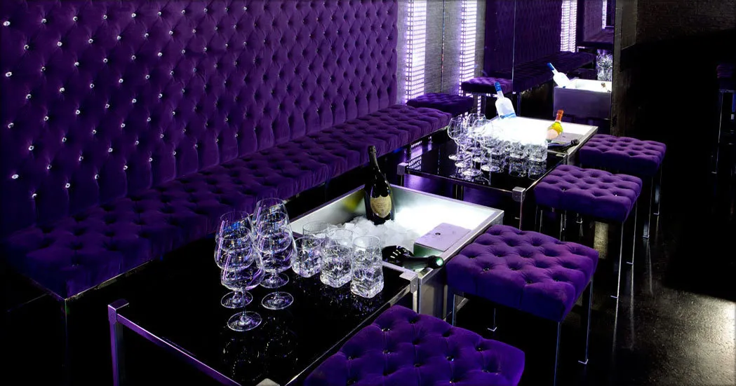 Espace VIP du Boomboom Club à Bordeaux avec banquettes et murs capitonnés en velours violet, tables noires, verres à champagne et bouteille, éclairage LED violet et cristaux de diamant.