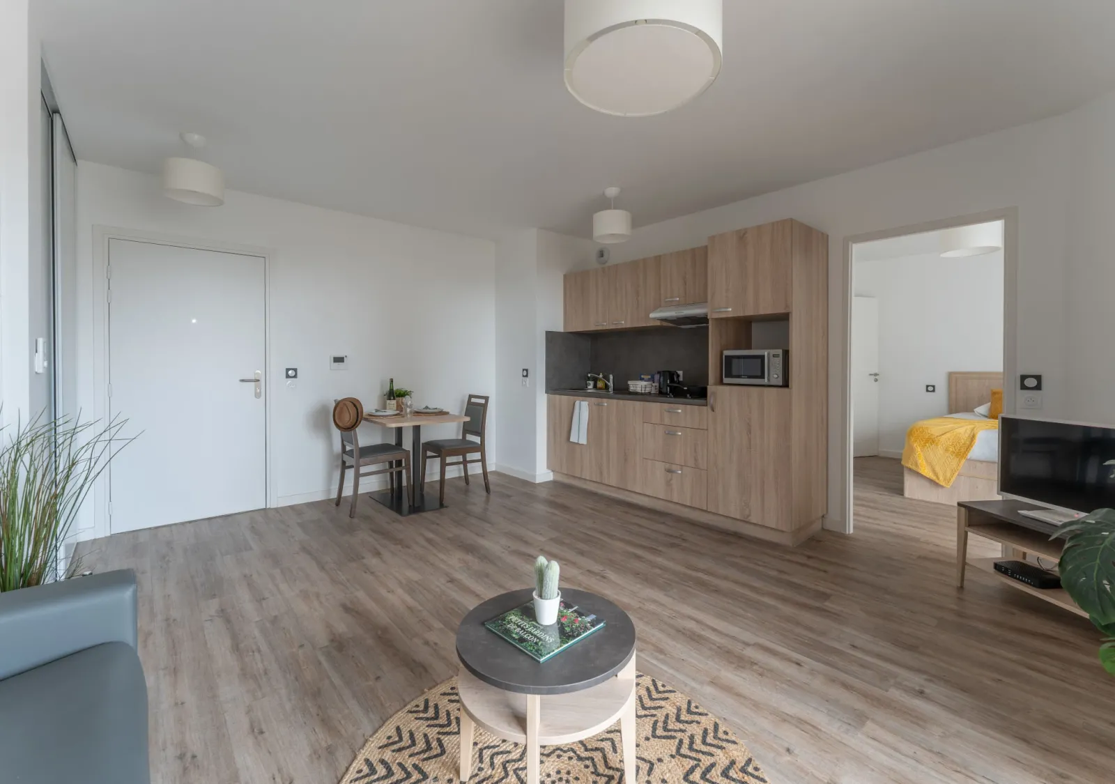 Studio moderne et lumineux avec cuisine équipée en bois clair, coin repas, salon avec télévision, chambre avec lit jaune, sol en vinyle imitation parquet, à la résidence Les Jardins d'Arcadie à Bordeaux.