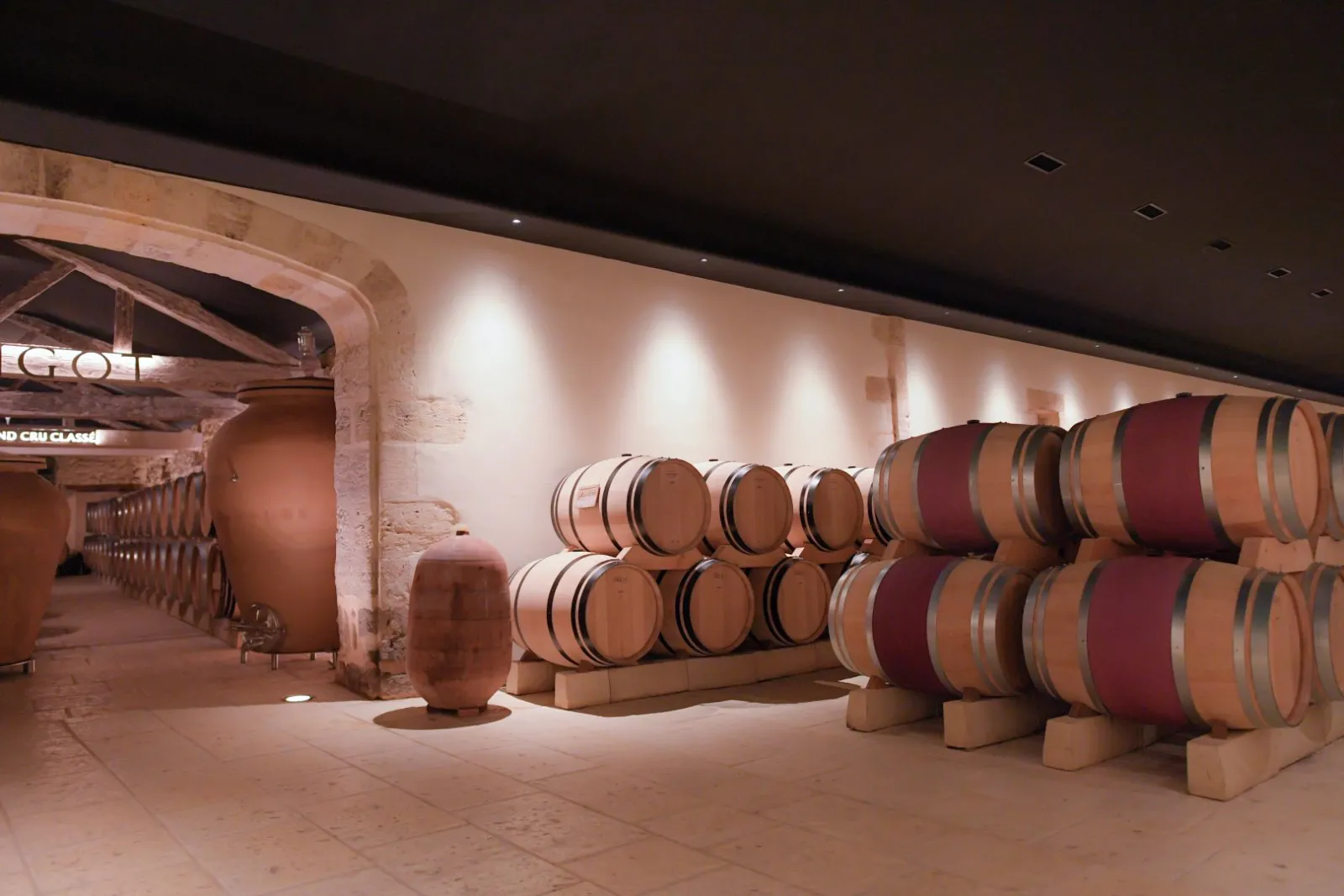 Cave de vinification du Château Mangot à Bordeaux avec des tonneaux de chêne alignés en rangées, illuminés par un éclairage moderne dans une architecture traditionnelle.