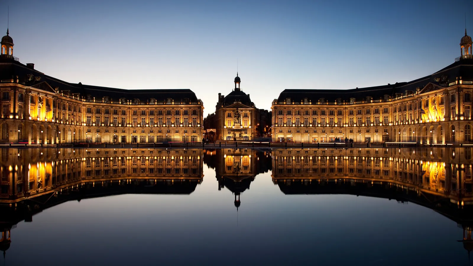 Place de la Bourse à Bordeaux illuminée la nuit, avec ses bâtiments classiques dorés symétriquement disposés autour d'un miroir d'eau reflétant les façades et le ciel crépusculaire.