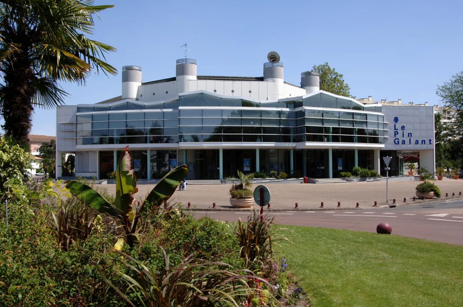 Bâtiment moderne du Pin Galant à Bordeaux avec façade en verre et acier, quatre cheminées blanches sur le toit, entouré de palmiers et d'espaces verts