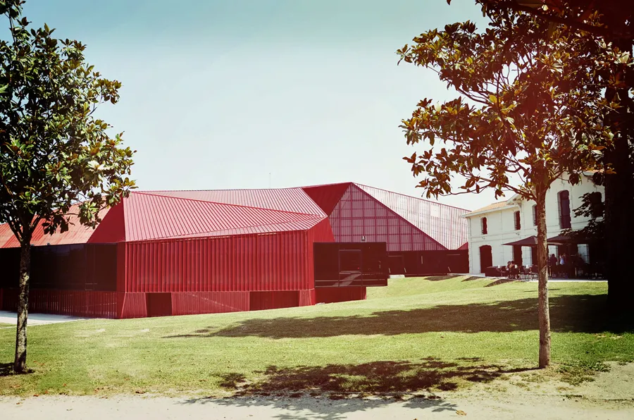 Vue du Rocher de Palmer à Cenon, avec un bâtiment contemporain aux façades ondulées en tôle rouge et blanche, entouré d'un parc arborisé et d'un bâtiment blanc traditionnel en arrière-plan.