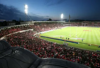 Vue aérienne du Stade Chaban-Delmas de Bordeaux rempli de spectateurs en tenue rouge et blanche lors d'un match de rugby en soirée, avec le terrain éclairé par des projecteurs.