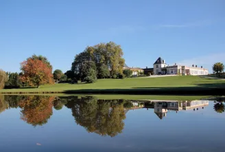 Vue du Château Paveil de Luze à Bordeaux avec sa façade blanche classique se reflétant dans l'étang, entouré d'arbres aux feuillages d'automne et d'une vaste pelouse verte sous un ciel bleu.