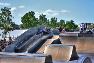 Vue du skate park de Bordeaux avec ses rampes en béton, des skateurs en action, des spectateurs, et la Garonne en arrière-plan sous un ciel bleu.