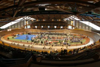 Vue panoramique du vélodrome de Bordeaux avec piste cycliste ovale, spectateurs en tribunes, et installations sportives intérieures sous structure métallique.