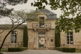 Façade élégante du château Cordeillan-Bages à Bordeaux, avec architecture en pierre de taille, balcon forgé, entrée principale ornementale, cyprès encadrant l'accès, et végétation environnante.