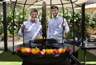 Deux chefs cuisiniers en tenue blanche posent devant un grill avec des tomates fraîches à la Terrasse de Lafaurie à Bordeaux, entourés de fleurs blanches et roses.