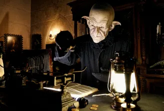 Créature fantastique maquillée portant un costume noir dans un décor gothique de Bordeaux, entourée de lanternes lumineuses et d'objets anciens.