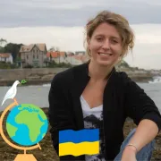 Femme souriante portant un t-shirt aux couleurs du drapeau ukrainien bleu et jaune, posant devant un paysage fluvial avec maisons anciennes. Un globe terrestre et une colombe de paix sont superposés à l'image.