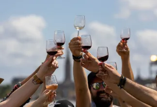 Groupe de personnes levant des verres de vin rouge et blanc en toast, avec la ville de Bordeaux en arrière-plan sous un ciel nuageux.
