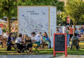 Groupe de personnes assises autour d'une grande pancarte intitulée 'Le vignoble de Bordeaux' lors d'un événement public en plein air à Bordeaux, avec un panneau de menu en premier plan.