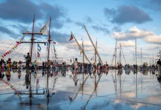 Plusieurs voiliers traditionnels amarrés dans une baie peu profonde à Bordeaux, avec des visiteurs se promenant sur l'eau reflet du ciel nuageux.