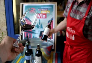 Un verre de vin rouge tenu à la main lors d'une dégustation à Bordeaux, avec des bouteilles et un présentoir promotionnel en arrière-plan