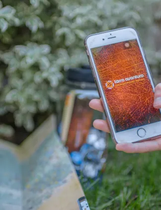 Main tenant un iPhone blanc affichant l'application Terra Aventura avec une carte orange, dans un jardin avec des plantes vertes et une carte papier en arrière-plan à Bordeaux
