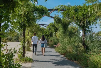 Deux personnes marchent dans une allée ombragée bordée de vignes et de végétation luxuriante, avec des structures en bois entrecroisées au-dessus, dans les jardins de Bordeaux.