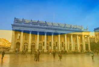 Le Grand Théâtre de Bordeaux avec ses colonnes classiques illuminées aux couleurs bleu et jaune en soutien à l'Ukraine, sous un ciel bleu dégagé avec des visiteurs dans la place.