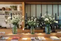 Table en bois avec trois vases en verre contenant des marguerites blanches et jaunes, suspendus au-dessus par des cordons noirs, dans une cuisine contemporaine avec étagères en arrière-plan et magazines éparpillés.