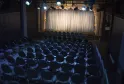 Salle de théâtre vide avec rangées de fauteuils noirs face à une scène éclairée avec rideau blanc et draperies dorées, à Bordeaux.